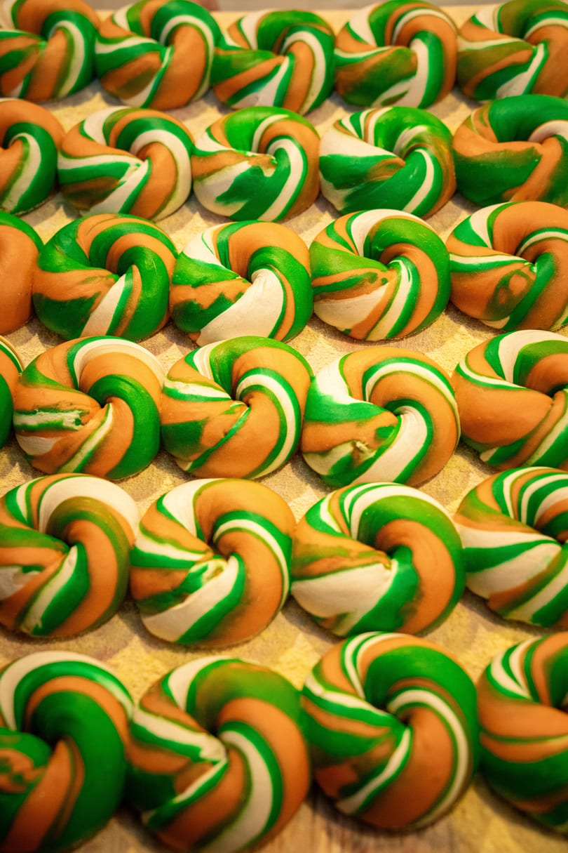 Rows of colorful green, white, and orange swirled bagels on a light-colored surface.