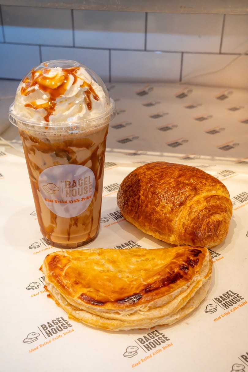 Iced coffee with whipped cream and caramel, a croissant, and a pastry on Bagel House branded paper.