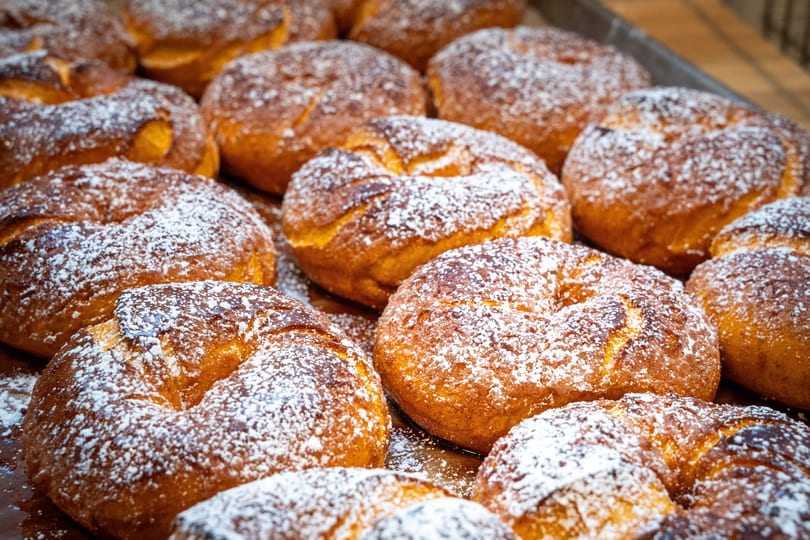 Close-up of many golden-brown, ring-shaped pastries dusted with powdered sugar on a tray.