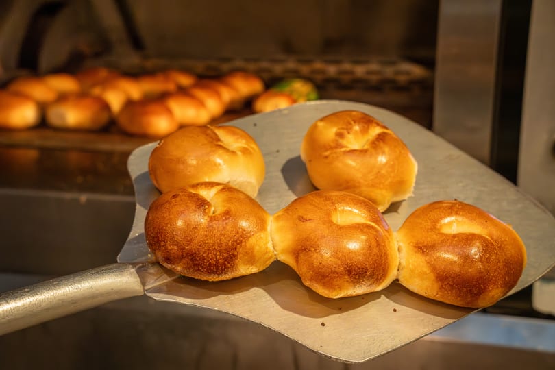 Freshly baked golden-brown bagels being removed from a hot oven with a metal peel.