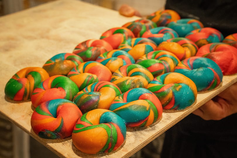 Many colorful rainbow bagels, swirled with red, orange, green, and blue, on a wooden tray.